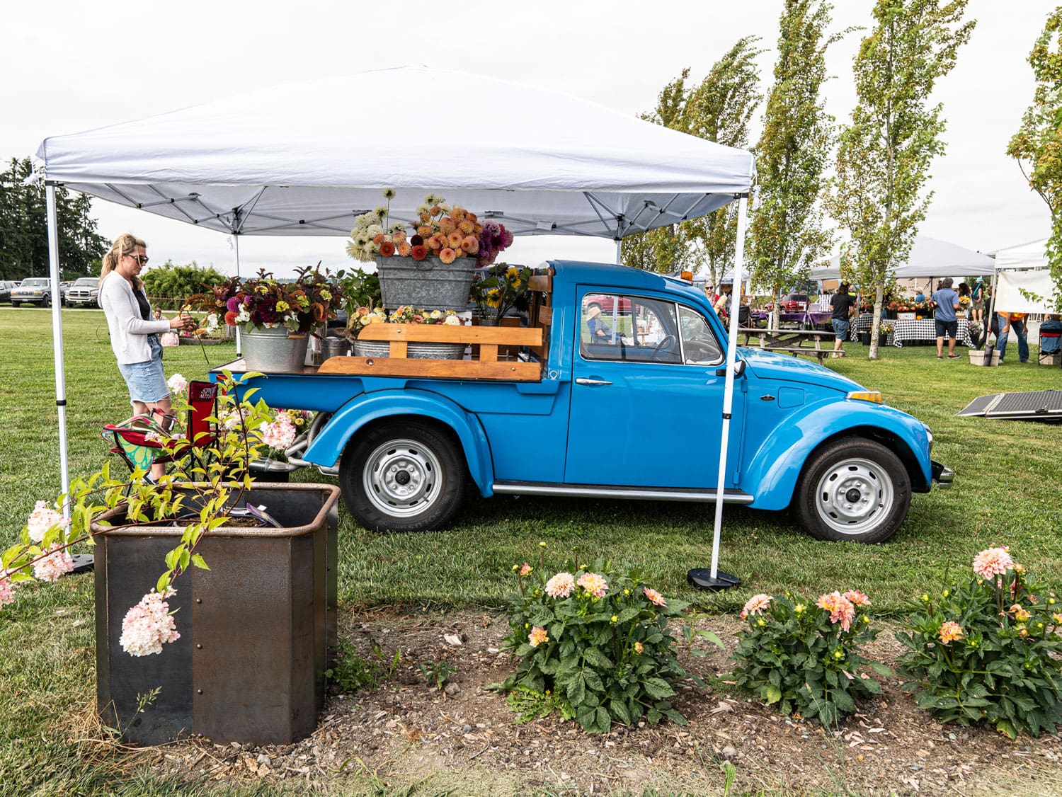 Vintage blue pickup truck filled with flower arrangements parked under a canopy at an outdoor market with vendors and shoppers nearby.