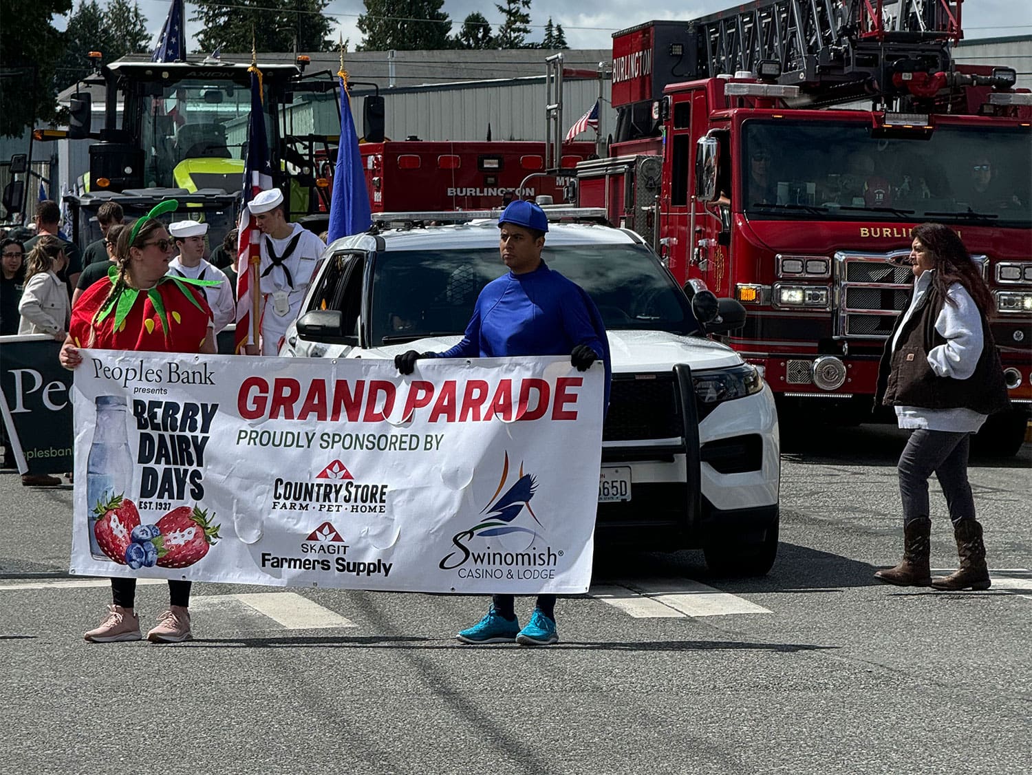 People carrying a “Berry Dairy Days Grand Parade” banner while walking in a small-town parade with a fire truck behind them.