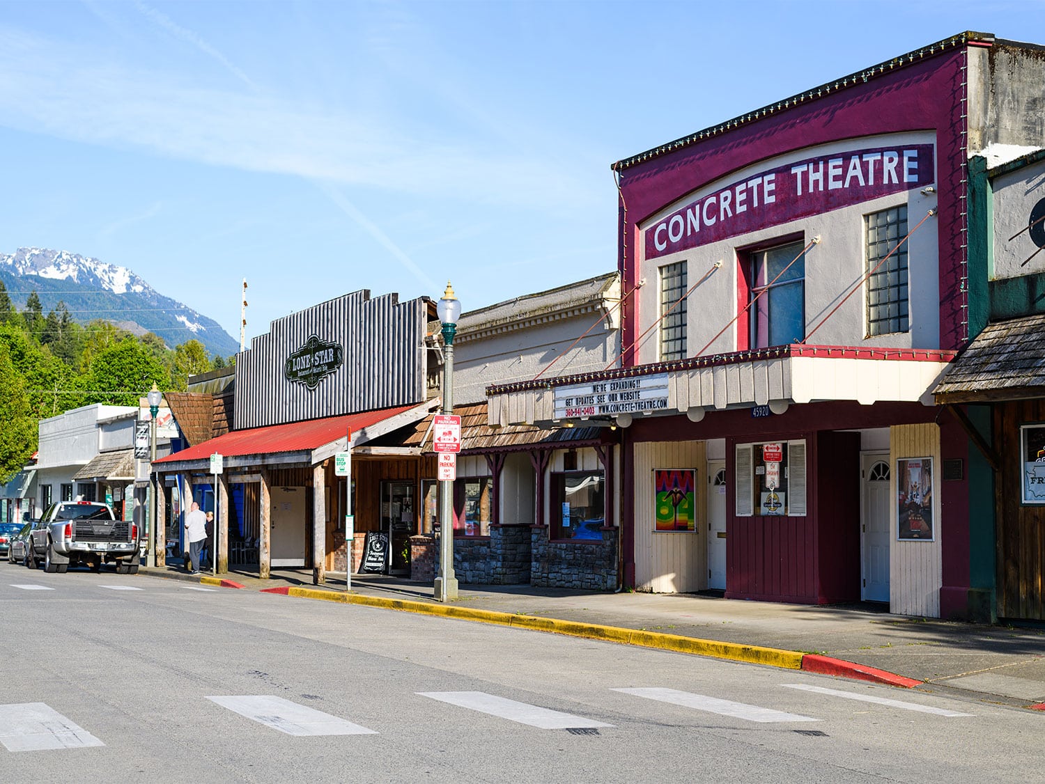 The charming main street of Concrete, Washington, featuring the historic Concrete Theatre and local shops, with snow-capped mountains rising in the background.