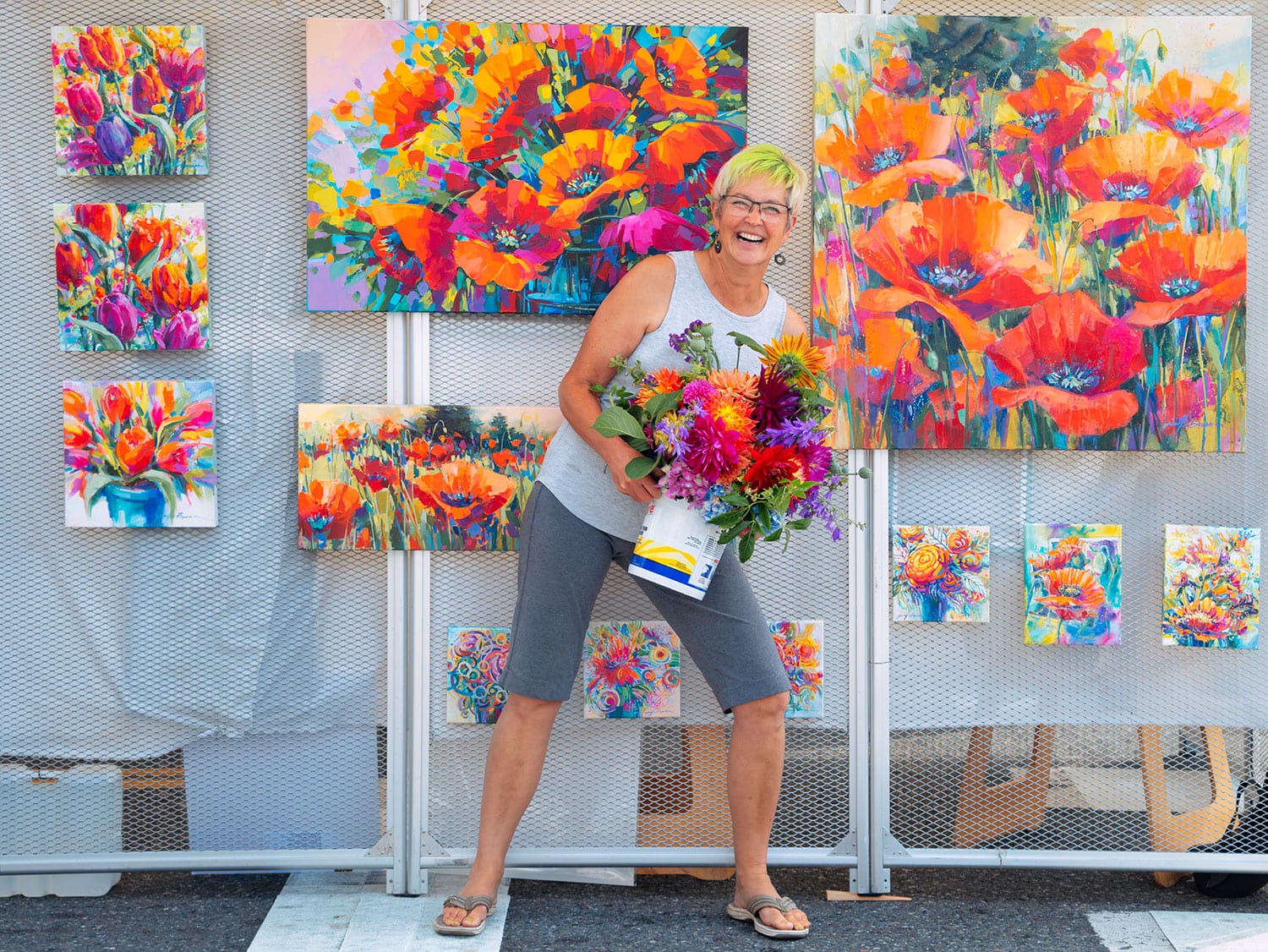A smiling artist holds a bouquet of colorful flowers in front of her vibrant floral paintings displayed at the Anacortes Arts Festival in Washington.