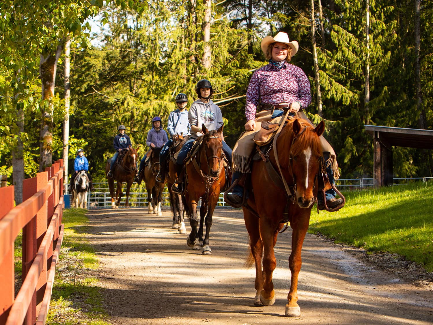 A group of horseback riders led by a smiling woman in a cowboy hat along a sunny tree-lined trail in Skagit County, Washington.