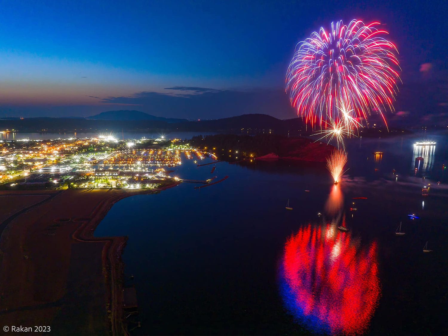 Aerial view of red, white, and blue fireworks bursting over the water and reflecting off the bay alongside a lit-up Skagit County waterfront community at dusk.