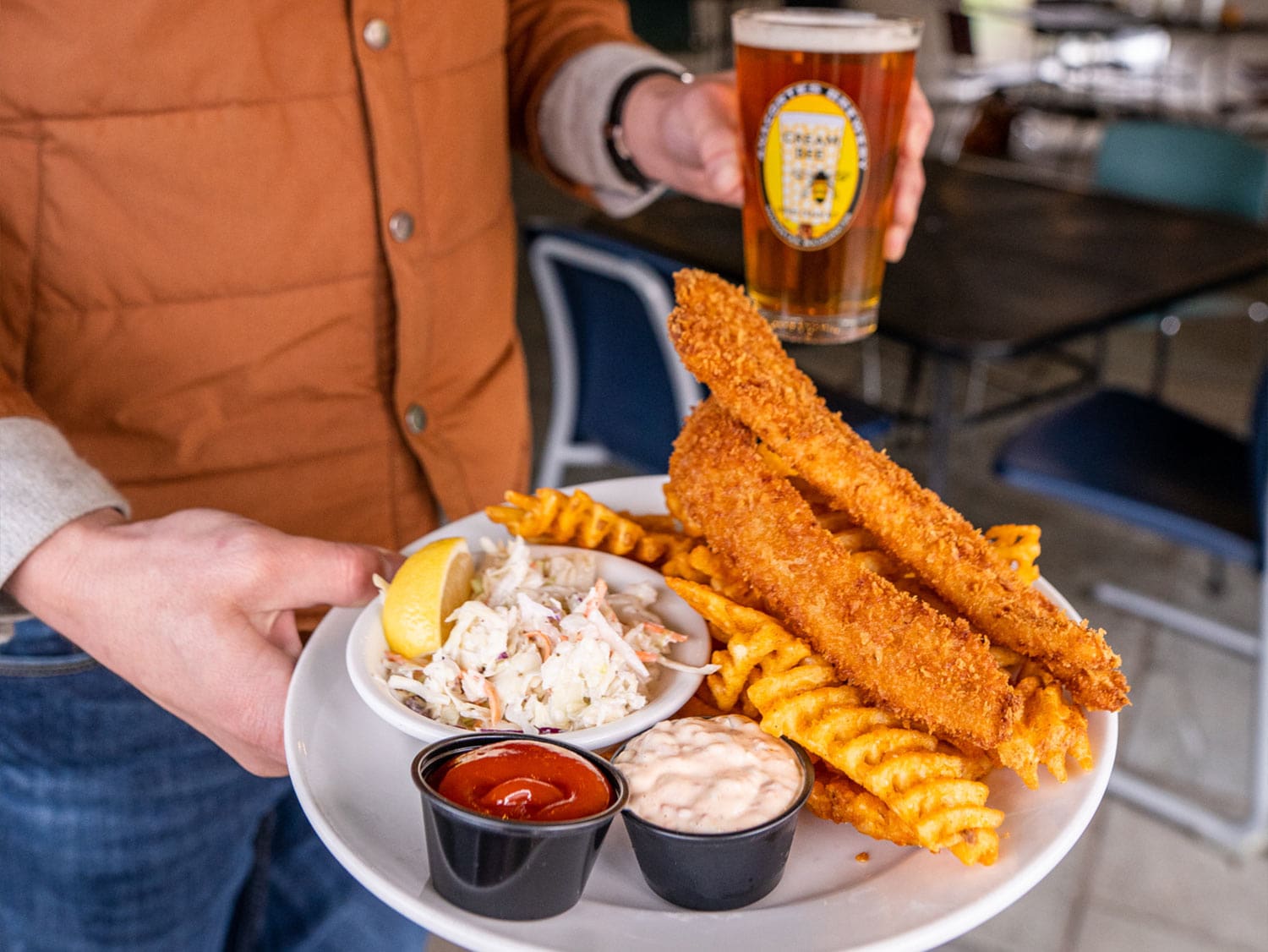 A person holds a plate of fish and chips with coleslaw and dipping sauces alongside a pint of craft beer at a Skagit Valley event.