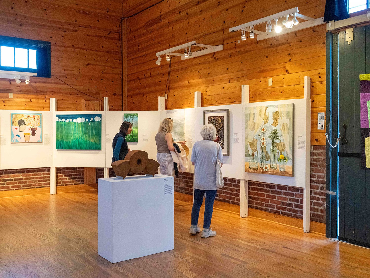 Visitors browse paintings and sculpture in a warmly lit art gallery with wood-paneled walls and exposed brick in Skagit Valley, Washington.